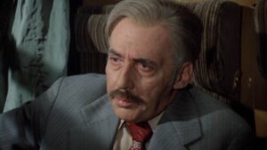 Older man with gray hair and mustache, wearing a gray suit and red tie, looking thoughtful inside a dimly lit cabin.