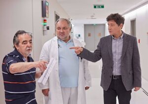 Three men in a hospital corridor gather around a document as they discuss plans, one in a striped polo, one in a white lab coat, the third in a gray blazer point to the paper.
