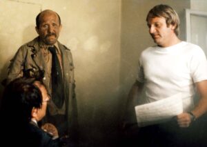 Two men in a room: a younger man in a white T-shirt holds papers while an older man in a tattered jacket looks on, with a blurred person in the foreground.
