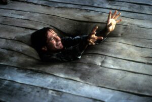 Man reaches upward from a circular hole in a weathered wooden floor, arms outstretched as he tries to climb out.