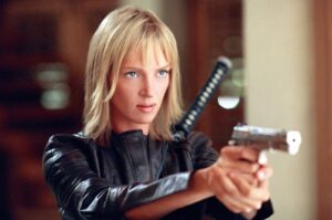 Blonde woman in a black leather jacket aiming a silver handgun with a katana strapped to her back.