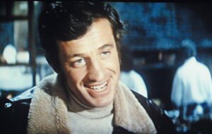 Candid close-up of a smiling man with dark hair, wearing a light-colored shearling-collared jacket, outdoors.