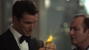 Man in a tuxedo holds a glowing gold nugget wrapped in foil while another man in a suit looks on in a dim background.