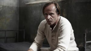Man with light hair in a beige knit sweater leaning on a metal bed frame in a dim, concrete room, looking at the camera.