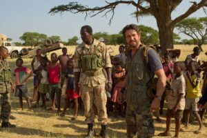 Soldiers in camouflage talking with local children in a dry savanna; an overturned vehicle lies behind them under a tree.