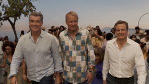 Three men in button-down shirts walk toward the camera at a seaside party with a cheering crowd behind them near the ocean and sunset, smiling.
