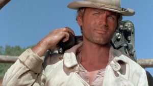 Man in a beige shirt and cowboy hat carries a guitar case over his shoulder outdoors against a blue sky.