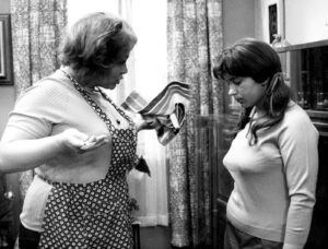 Older woman in an apron confronts a pregnant younger woman in a kitchen setting.