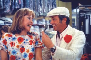 A man in a light checkered suit and white cap shares a laugh with a woman in a bright floral dress inside a shop, suggesting a playful moment.