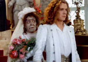 A pale, zombie-like bride in a wedding veil stands behind a young man in a white suit and bouquet in a formal room.
