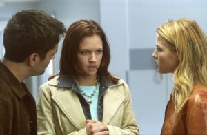 Three young adults in a hallway having a tense conversation; center woman with brown hair wearing a denim jacket and beige coat, flanked by a man and a blonde woman in leather jacket.