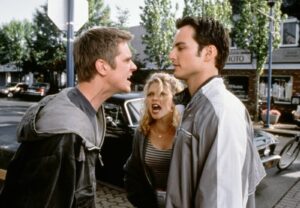 Two men face off in a street confrontation while a blonde woman angrily shouts between them, by a parked car and storefronts in the background.