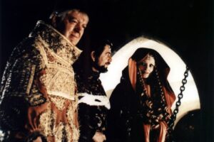 Three people in elaborate medieval costumes stand together in a dark setting, with a bearded man in a gold-embellished robe, a second man, and a woman in a red hooded cloak with braids.
