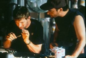 Two men in a workshop focus on a small task; one man uses a tool while the other observes nearby at a cluttered workbench.