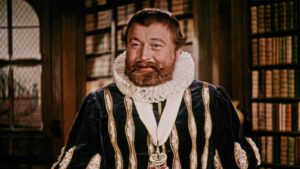 Man with a beard wearing a dark velvet outfit and a large white ruff, standing in a library.