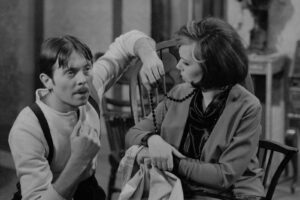 Vintage photo of a man in a sweater and suspenders talking to a woman who holds a bead necklace; they sit indoors.