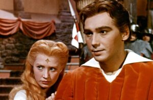 Man in a red tunic with white collar stands in foreground, beside a woman with long red hair and a star-shaped mark on her forehead, in a medieval/fantasy setting.