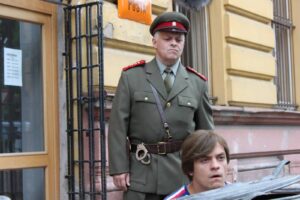 Male officer in green military uniform with red shoulder boards, standing outside a yellow-brick building; handcuffs hang from his belt while a younger man sits nearby.