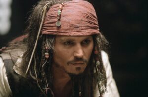 Close-up of a man with long hair wearing a worn red bandana and beaded accessories, in pirate attire, on a dark background.