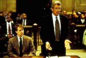 Attorney in a dark suit stands and speaks at a bench; a seated colleague in a tan suit sits nearby.