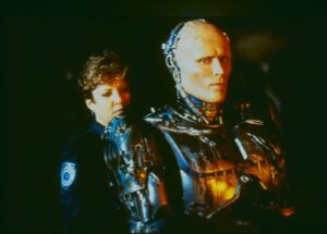 RoboCop-like figure in a metallic suit with a female officer in the dimly lit background.