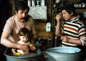 A man and a younger man help bathe a small child in a plastic tub, using water toys in a rustic workshop/kitchen setting with tools in the background.
