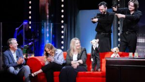 Five people sit on a bright red couch on a TV talk-show set, laughing and chatting while two puppets stand nearby on the table stage right.