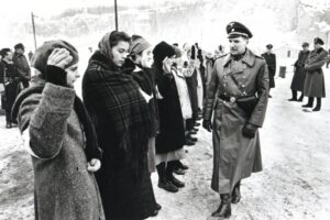 Uniformed officer walking past a line of women and men in winter coats during a cold outdoor inspection. Snowy ground in the background.