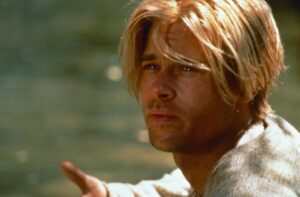 Blond-haired man with stubble outdoors, looking intently and pointing forward with his hand extended.