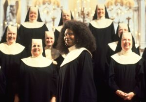 African American woman in a choir robe stands in front of a group of nuns in a church setting.
