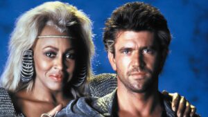 Two adults pose closely for a portrait: a woman with platinum hair and large hoop earrings beside a man with stubble against a blue background.