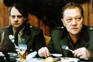 Two men in green military uniforms sit at a dining table with plates, bread rolls, and a tall glass of beer in front of them.