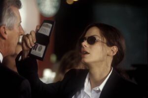 Woman in a blazer with sunglasses presents or shows a badge to a man holding a handheld badge scanner or ID reader in a dim setting.