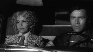 Black-and-white photo of a man driving and a woman beside him in the car, both looking ahead with serious expressions.