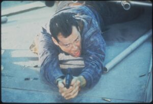 Man in a blue plaid shirt on his hands and knees on pavement, reaching forward with a tense expression.