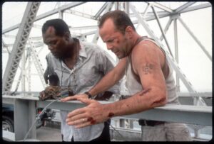 Two construction workers lean over a metal railing, showing bloodied arms from a recent injury on a scaffold-like structure.