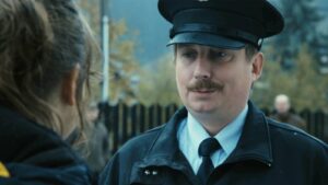 Police officer in uniform and cap talking to a person outdoors, with a fence and trees in the background