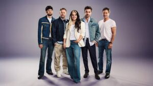 Group of five adults posing together in a studio; everyone wears casual denim or smart-casual outfits against a purple-gray backdrop.