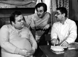 Three men in a cluttered room discuss notes at a table: a heavyset man seated left, a man leaning on a chair in the middle, and a man with glasses and a white shirt pointing to papers on the table (black-and-white photo).