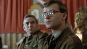 Two soldiers in vintage uniforms sit indoors; foreground man with round glasses gazes to the right, another soldier behind him looks forward.