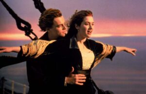 Couple with arms outstretched on a ship's bow at sunset, embracing and looking ahead over the ocean.