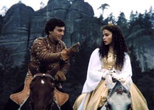 Two costumed riders on horseback outdoors in a forested, rocky setting: a man in brown studded armor points while the woman in a cream gown with a small crown looks on.