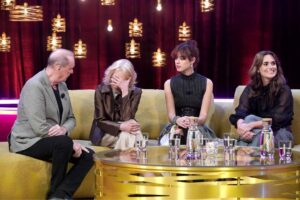 Four people seated on a gold sofa during a panel discussion, with a glass coffee table and water bottles in front; warm pendant lights and red curtains in the background.