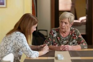 Two women sit across from each other at a table, conversing; the older woman wears a floral blouse.