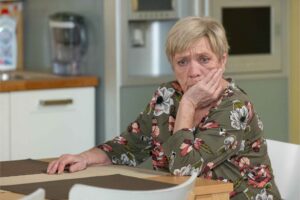 An older woman in a floral dress sits at a table, resting her hand on her cheek with a worried expression in a kitchen setting.