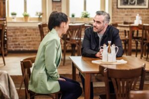 Two adults sit across from each other at a wooden table in a cozy cafe, mid-conversation.