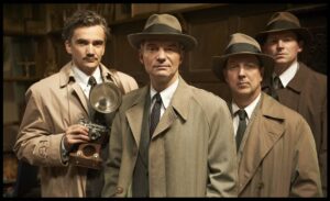 Four men in 1940s-style trench coats and fedoras pose indoors, one holding a vintage film camera with a large flash