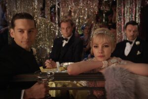 Man in tuxedo and woman in flapper-style gown lean on a gilded railing at a glamorous party, holding champagne glasses.