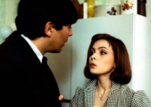 Woman with short brown hair and a checkered blazer speaks to a man in a suit in a kitchen setting.