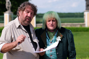 Two adults outdoors; man in a beige shirt holds papers and a pen, while a woman with green hair stands beside him.